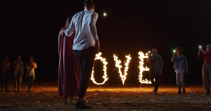 Young Man Surprising his Girlfriend with Marriage Proposal With Big Letters of the Word LOVE Burning in Pyrotechnic Fire. Friends Filming and Cheering as Woman Accepts to Marry her Boyfriend
