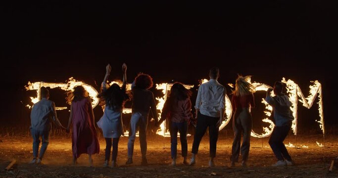 Cinematic shot of Young, Carefree, Happy multi-ethnic Friends Having fun, Running Hand in Hand Towards Burning Letters Spelling the Word FREEDOM. Group of Young People Dancing in Back View Shot 