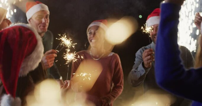 Cinematic shot of Young Happy Smiling Multi-ethnic Friends with Santa Hats and Sparklers in their Hands Singing Christmas Songs and Dancing Together During Celebration Party Outdoors at Night.