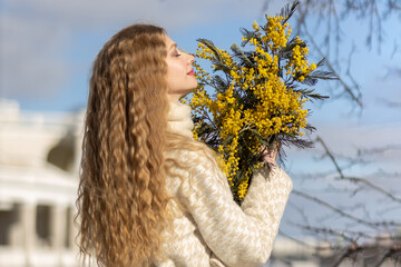 A young woman stands with a bouquet of yellow mimosa and sniffs the flowers. The concept of the Spring holiday - March 8, Easter, Women's Day