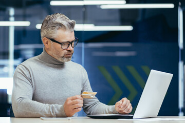 Senior mature businessman inside the office holds a bank credit card in his hands, gray-haired boss is serious and thoughtful makes a money transfer and purchase in an online store, online booking.
