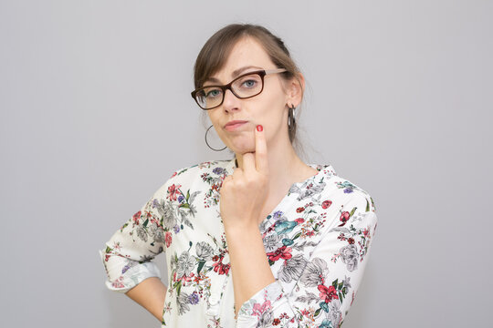 Young woman with glasses holding her finger on her face and wondering about something. She has brown hair, white floral blouse and big oval earrings. Photo on plain grey background