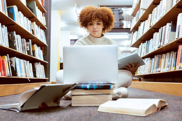 College library learning, laptop and black woman student working on the floor with books. Reading,...