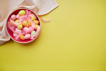 Minimalist Easter bowl on towel with colorful eggs and pink feathers on yellow backgound. Place for text.