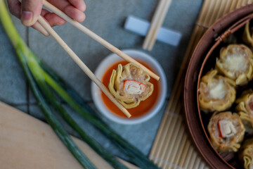 Dim sum with chopsticks and top view over a bowl of chili sauce and a portion of dimsum on in steamed bamboo on a bamboo mat and a spring onion. Top View