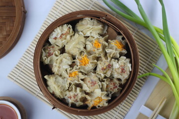a portion of dimsum in steamed bamboo on a bamboo mat and surrounded by sauce and a piece of green onion. White background. Top view