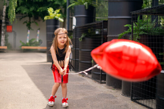 A little hippie girl in a red leather skirt holds a balloon in the shape of a star. A child walks in the city courtyard