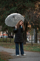 Young woman using smartphone under umbrella. Woman with transparent umbrella walking in park in fall and talkin to phone