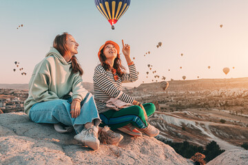 Girl friends sitting on a clifftop viewpoint and admiring view of majestic flying hot air balloons...