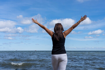 Beautiful woman raising her arms on the beach. She is dressed in a black T-shirt and white pants.