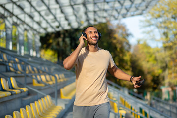 Young man walking in stadium after exercise and jogging in headphones, listening to audiobook music and online radio podcast, dancing joyfully, hispanic man on jogging day.