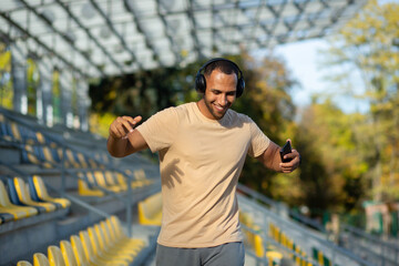Young man walking in stadium after exercise and jogging in headphones, listening to audiobook music and online radio podcast, dancing joyfully, hispanic man on jogging day.