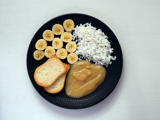 Banana slices, boiled rice, applesauce and toast on a black plate.