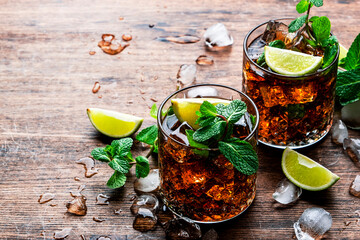 Rum cola cocktail with strong alcohol and ice, garnished with mint and lime in glass. Wooden background, hard light