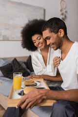 happy african american man holding tray with pancakes and drinks near sexy girlfriend in bedroom