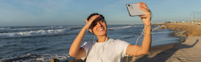 cheerful woman in wired earphones taking selfie near sea in Spain, banner.