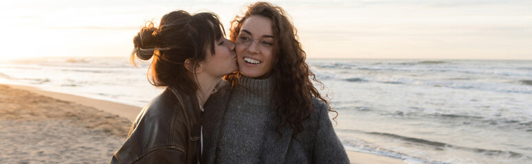Young woman kissing cheeks of cheerful friend on beach in Spain, banner.