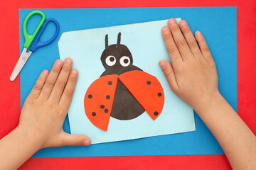 Top view of child hands with papercraft ladybug made by child and scissors on multicolor sheets background © Tatiana Foxy