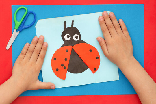 Top view of child hands with papercraft ladybug made by child and scissors on multicolor sheets background