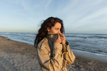 curly young woman covering face with collar of beige trench coat on beach in Barcelona.