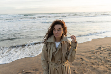 Young woman in trench coat touching curly hair on beach.