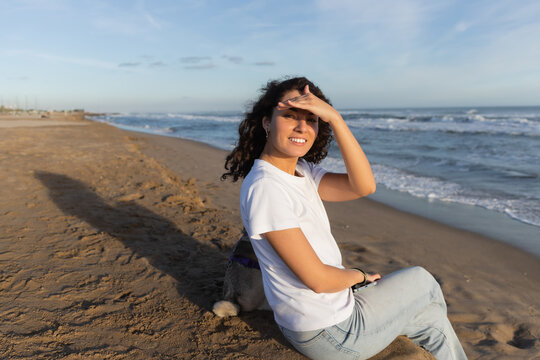 Cheerful Woman In White T-shirt Holding Smartphone And Sitting With Pug Dog On Beach In Barcelona.