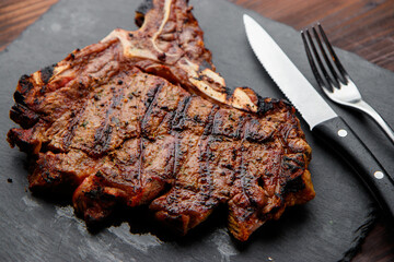 top view of delicious ribeye beef steak on the ribbon on the black stone plate with steel fork and knife in the wooden backgrounds 