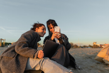 Smiling friends using smartphone and laptop on beach in Spain.