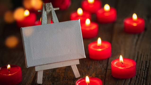 Blank canvas on an easle in front of many little red candlelights on rustic wooden underground - card for birthday, wedding, valentines day