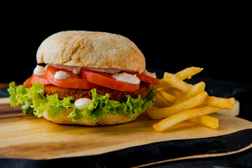 Classic hamburger with pork cutlet, tomato, salad leaves, white sauce with french fries on the wooden plate on the dark background   