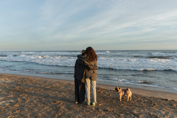 Back view of young women hugging near pug dog on beach in Spain.