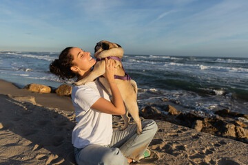 positive young woman with closed eyes holding pug dog on beach near sea in Barcelona.