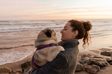 Side view of smiling curly woman in coat holding pug dog on beach in Barcelona.