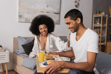 Obraz premium happy african american man holding tray with pancakes and drinks near smiling brunette woman in silk robe