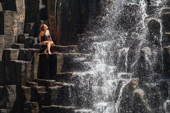 Caucasian woman in black swimsuit enjoying the falling water streams flowing on black volcanic stone cascades. Rochester Falls waterfall - popular tourist spot in Savanne district in Mauritius.