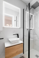 Bathroom with modern designer renovation and fittings. Vanity sink with black faucet. Walls of white granite with gray stains are beautifully illuminated by bright white light from square mirror.