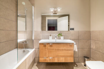 Bathroom in beautiful beige color. Vanity top sink with wooden drawers. Bathtub is fenced with...