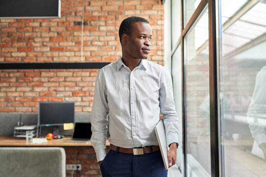 Thoughtful businessman in small startup office standing with laptop looking out the window