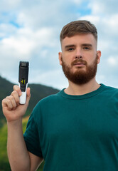 Handsome young bearded guy, European brutal man with a beard hair is shaving, trimming beard on face with a trimmer. Male personal self care, products, accessory on nature. Vertical photo.