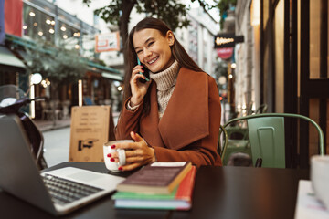 Smiling young woman using laptop and and talking on the phone while sitting in cafe