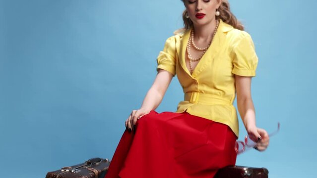 Hot day, travelling. Beautiful stylish young girl in yellow shirt and red skirt sitting on suitcases over blue studio background. Concept of retro fashion, beauty, attraction, 50s, 60s. Pin-up style