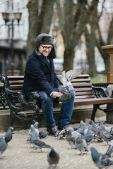 An elderly man enjoys a cold winter morning in the city. A gray-haired man feeds pigeons while sitting on a bench in the square.