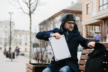 Senior freelancer or manager working on laptop while sitting on bench in city while on business trip. The concept of work on the go.