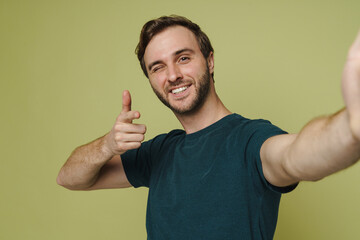 Smiling man winking and pointing finger at camera while taking selfie isolated over green wall