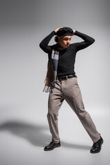 full length of young and trendy african american man in black pullover and plaid scarf posing with hands above head on grey background