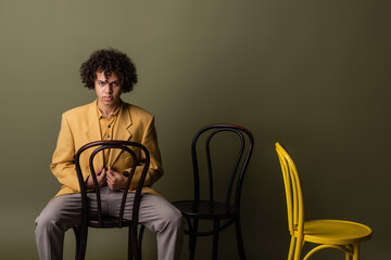 african american guy in stylish outfit posing with black and yellow chairs on olive grey background