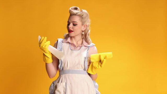 Beautiful stylish girl, housewife in retro apron and rubber gloves doing cleaning over yellow studio background. Concept of retro fashion, beauty, attraction, 50s, 60s. Pin-up style. Vintage