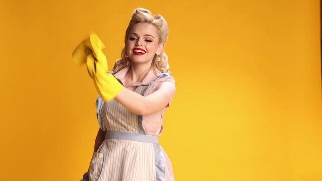 Smiling. Beautiful stylish girl, housewife in retro apron, rubber gloves doing cleaning over yellow studio background. Concept of retro fashion, beauty, attraction, 50s, 60s. Pin-up style. Vintage