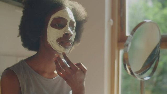 Medium close up shot of young African American woman applying clay mask to facial skin and looking in tabletop mirror during beauty routine at home