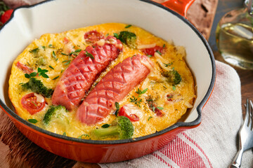 Omelette or frittata with two sausages, melted cheese, cherry tomatoes, broccoli and parsley on red skillet or iron pan on old wooden rustic table background. Healthy breakfast food concept. Top view
